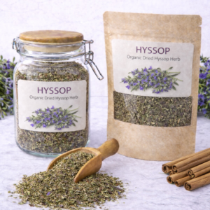 Organic dried hyssop herb in a glass jar and kraft resealable pouch with wooden scoop spilling loose cut leaves on marble surface, fresh purple hyssop flowers arranged around packaging.