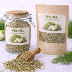 Organic fennel seeds in a glass jar and kraft resealable pouch with wooden scoop spilling loose fennel seeds on marble surface, fresh fennel bulbs and green fronds arranged around packaging.