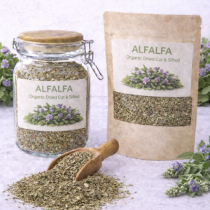 Organic dried alfalfa leaf cut and sifted in a glass jar and kraft resealable pouch with wooden scoop spilling loose alfalfa on marble surface, fresh alfalfa sprigs with purple blossoms arranged around packaging.