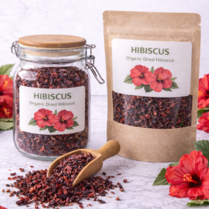 Clear glass jar and kraft paper pouch filled with organic dried hibiscus petals, surrounded by fresh red hibiscus flowers and a wooden scoop spilling loose hibiscus on a marble surface.