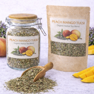 Peach Mango Tulsi herbal tea blend in a glass jar and kraft resealable pouch with wooden scoop spilling loose leaf blend on marble surface, fresh peach, mango slices, and tulsi flowers arranged around packaging.